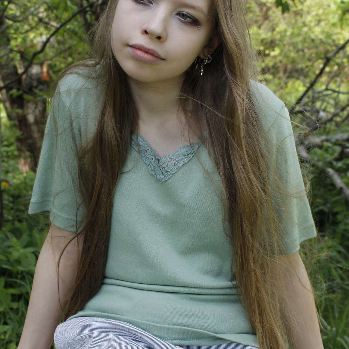 Woman sitting in a forest wearing a light green shirt and gray pants.