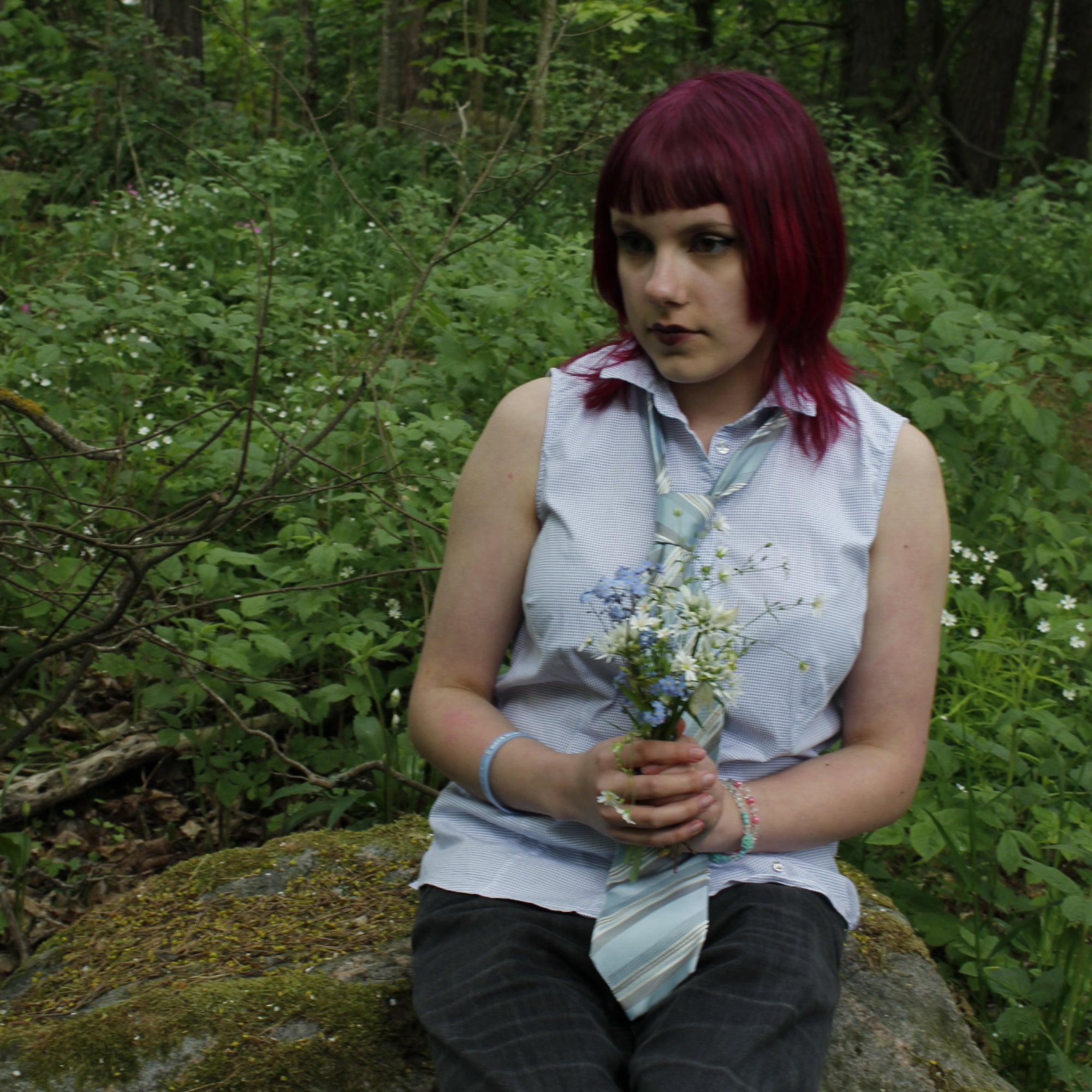 Person with red hair sitting on a rock in a forest holding flowers.